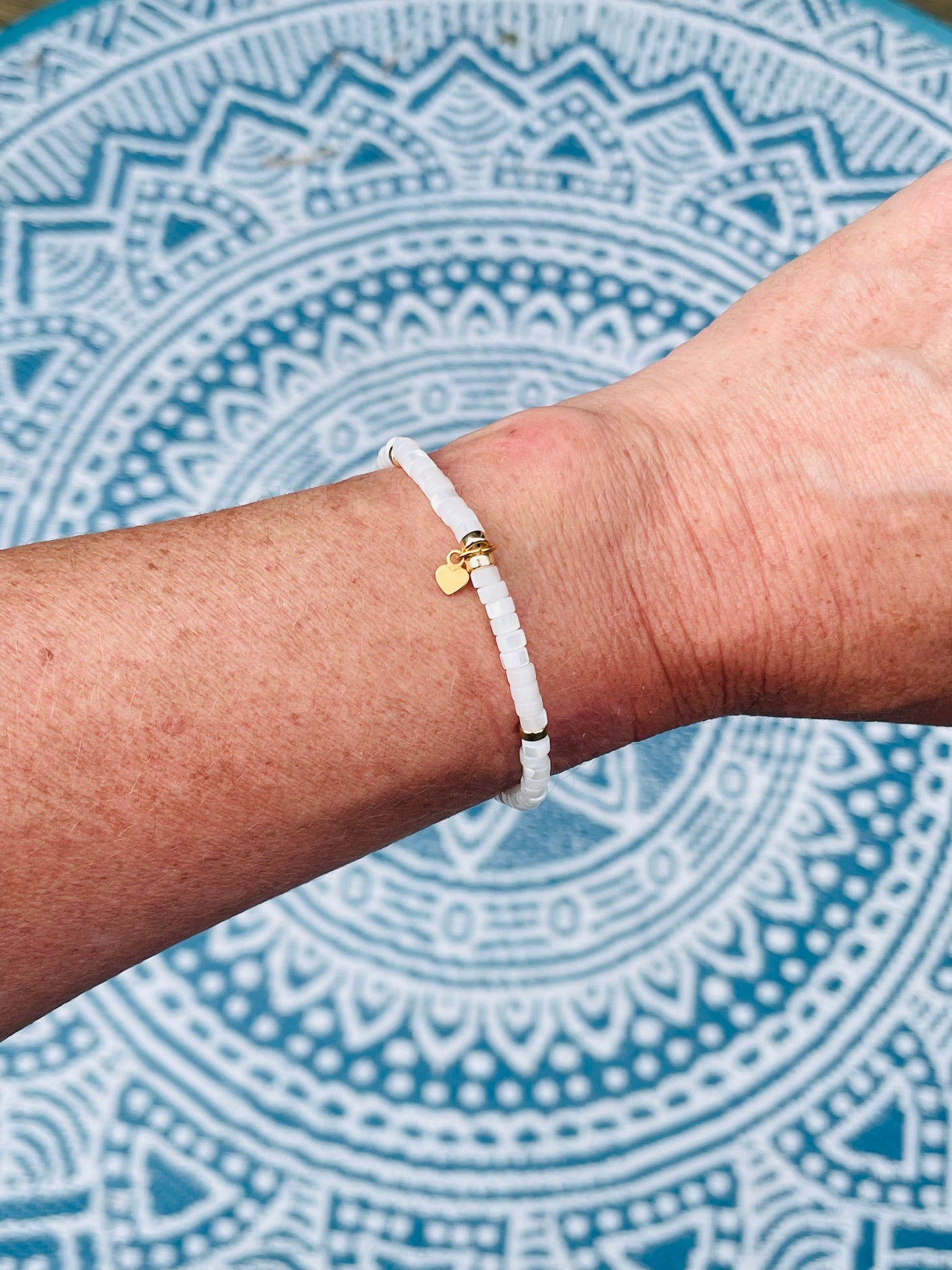 Mother of pearl and gold heishi bead bracelet with gold filled heart charm worn on wrist with mandala background