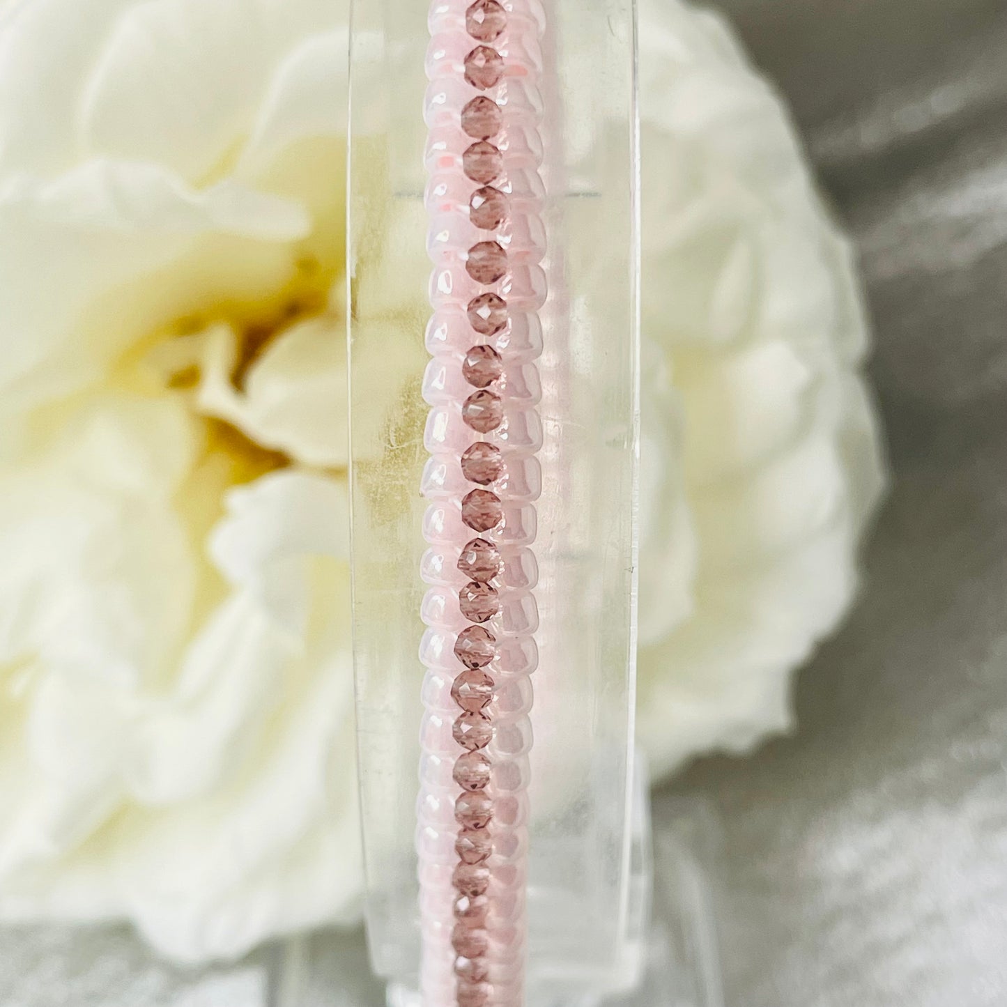 Close up of seed bead and crystal bracelet on clear stand with white rose in background