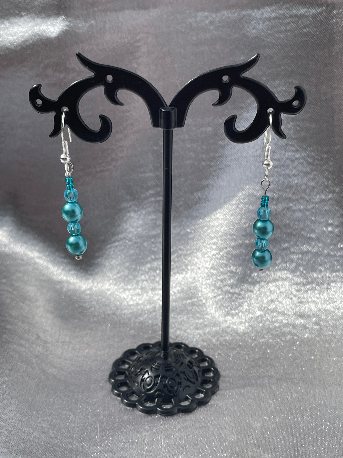 Crystal and pearl teal drop earrings on black stand with silver satin background