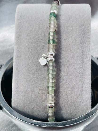 Close up of Fluorite and silver heishi bead bracelet with small heart charm on grey velvet stand with silver satin background