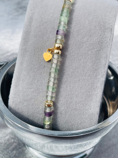 Close up of Fluorite and gold heishi bead bracelet with small heart charm on grey velvet stand with silver satin background