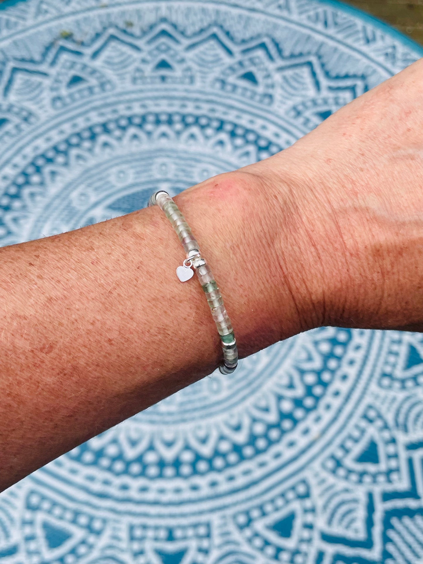 Fluorite and silver heishi bead bracelet with small heart charm on wrist with mandala background