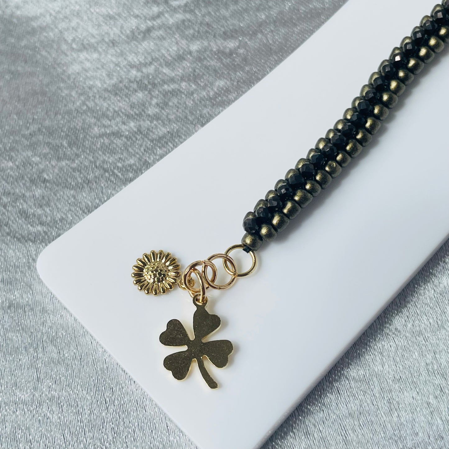 Bronze seed and black crystal bead bracelet with clover and daisy chases on white stand with silver background