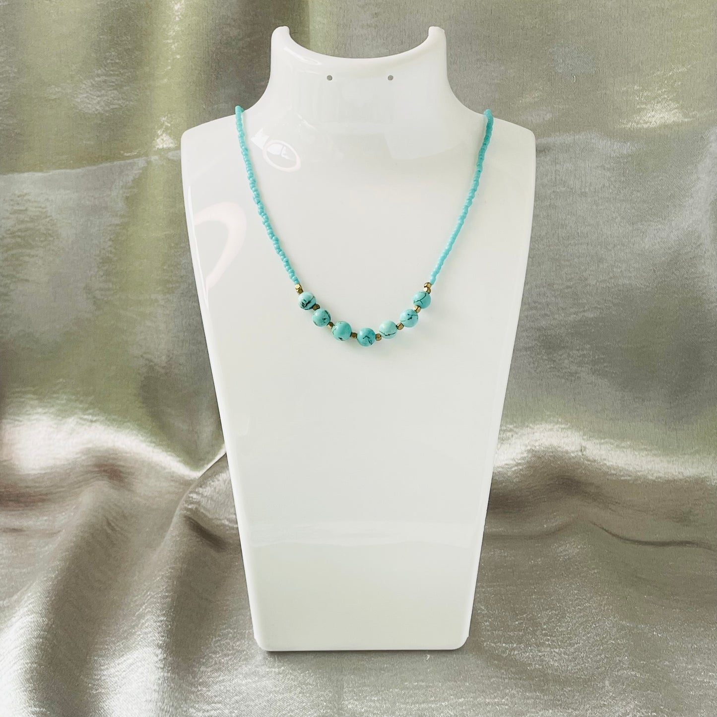 Turquoise seed and natural stone bead necklace with brass spacer beads on a white stand with silver background