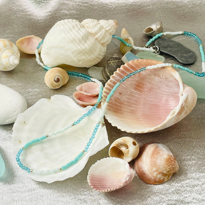 Seed bead and crystal necklace draped over seashells with silver background