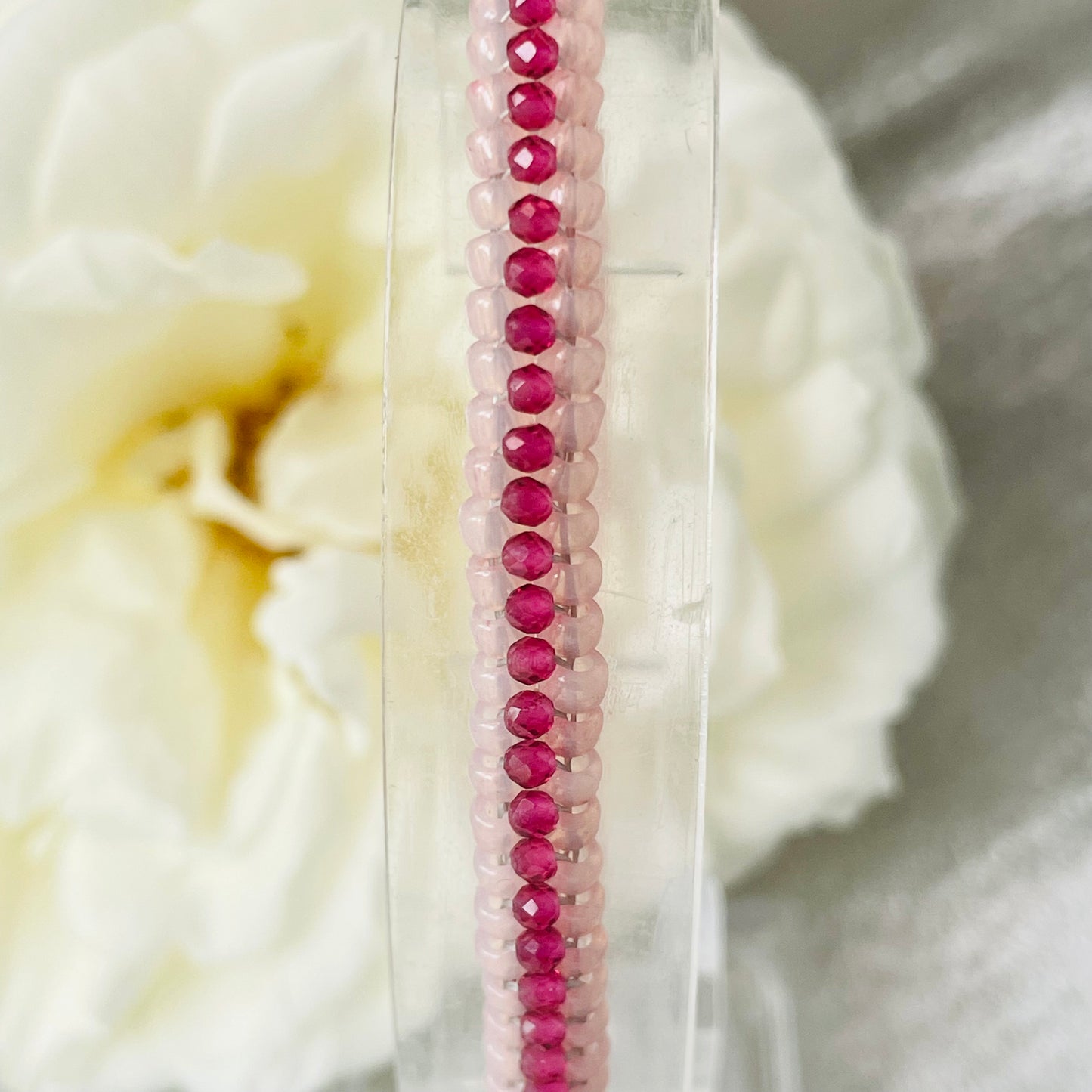 Close up of seed bead and crystal bracelet on clear stand with white rose in background