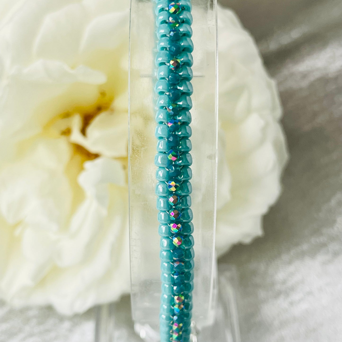 Close up of seed bead and crystal bracelet on clear stand with white rose in background