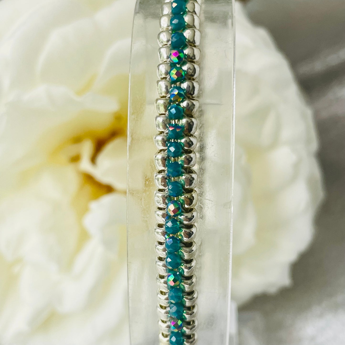 Close up of seed bead and crystal bracelet on clear stand with white rose in background